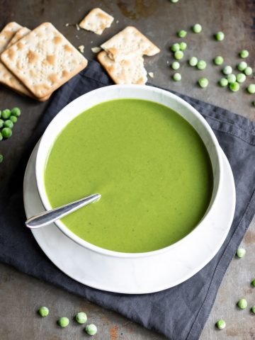A bowl of easy frozen pea soup next to frozen peas and crackers