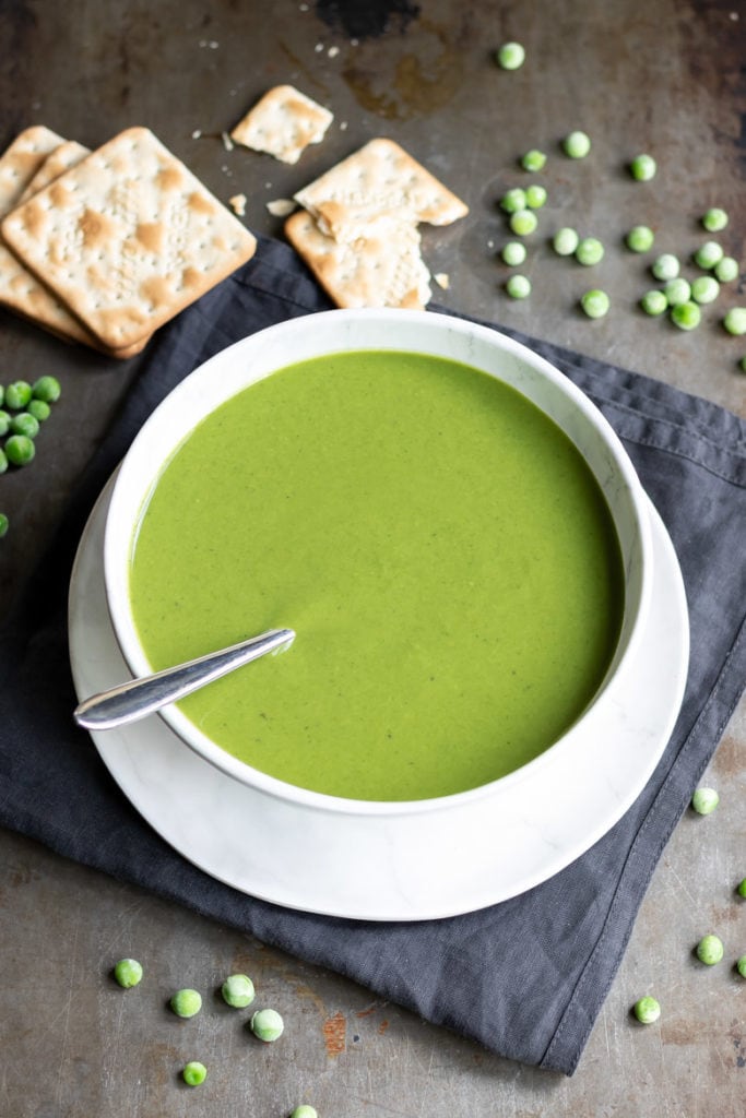 A bowl of easy frozen pea soup next to frozen peas and crackers.
