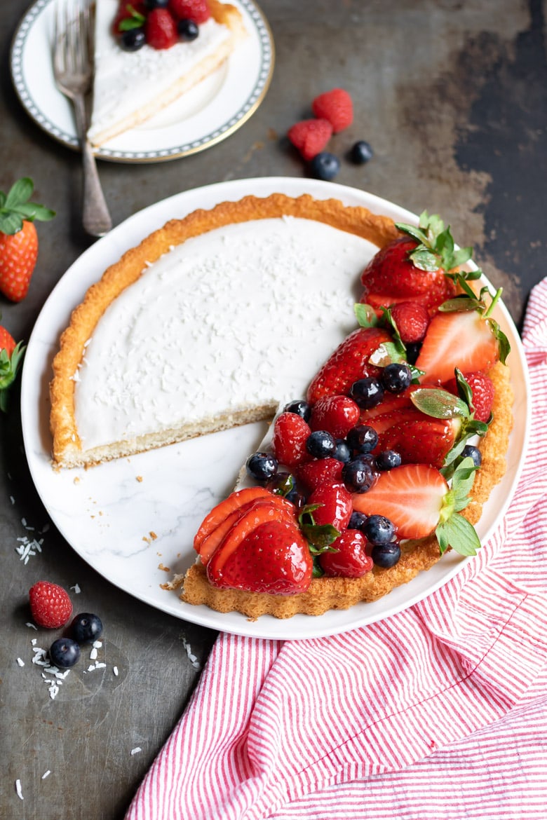 A Coconut Fruit Flan piled with glazed berries with a slice cut out and on a plate behind it.