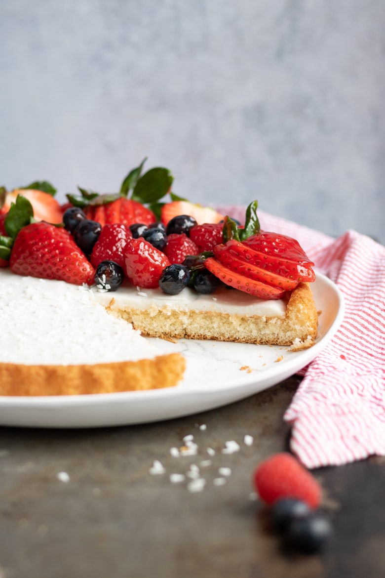 Side view of a Coconut Fruit Flan with a slice cut out. It has a sponge cake crust, creamy filling and is piled with glazed berries.