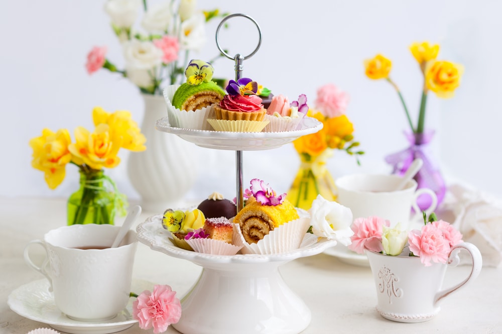 A cake stand full of food for afternoon tea, with tea cups and flowers.