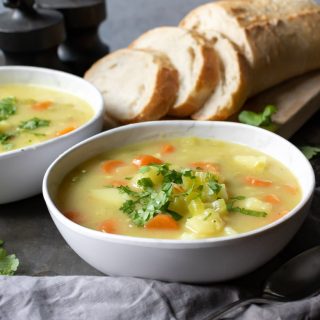 A close up of a bowl of vegan stew full of vegetables and potato, with slices of fresh bread in the background.