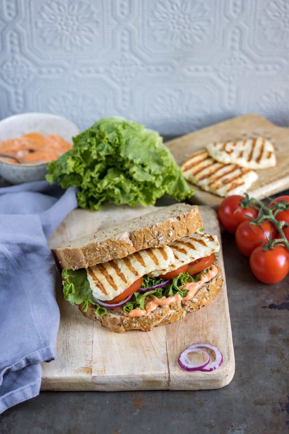 Table with a sandwich on a wooden board with lettuce, tomatoes and onion around it.