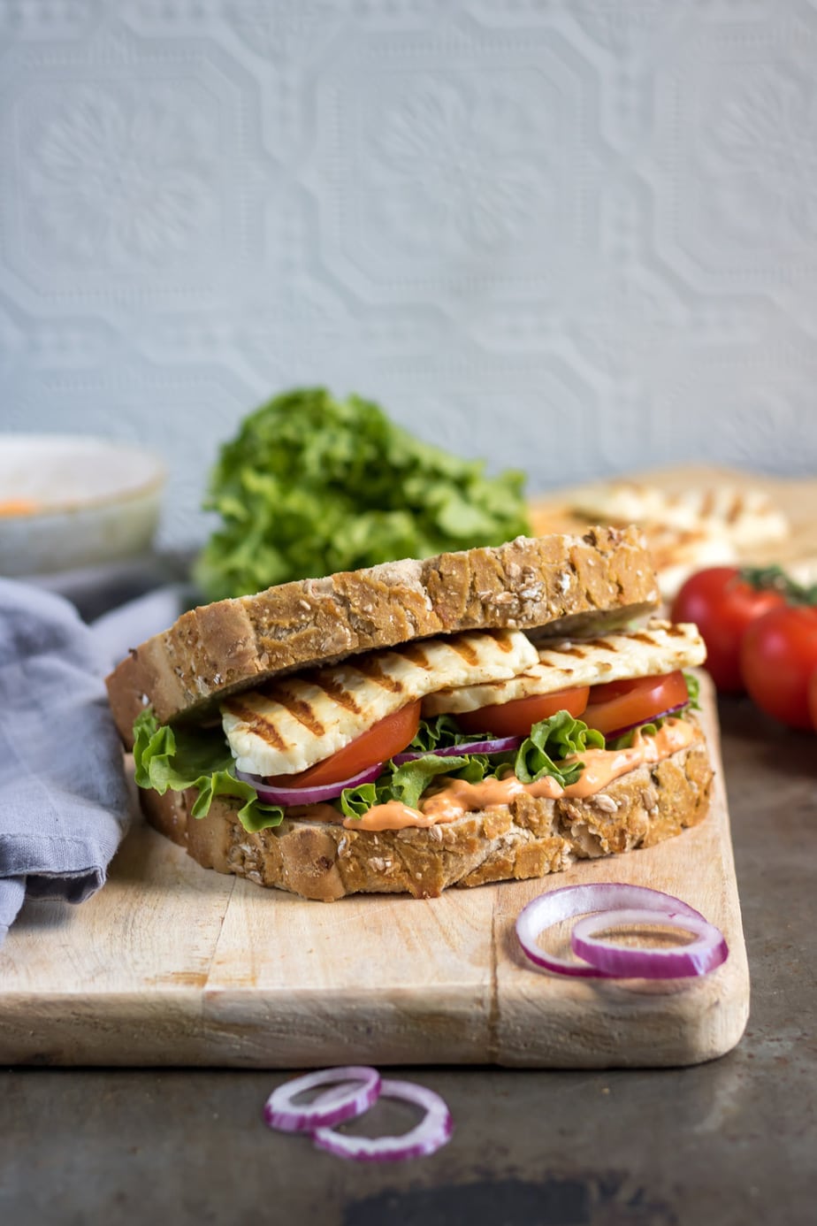 Close up of sandwich with halloumi, tomatoes, lettuce and sriracha mayo on a wooden board.