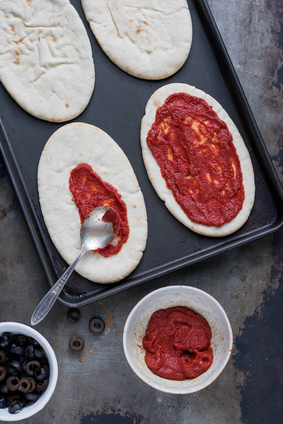 Pita breads on a baking sheet being spread with pizza sauce.