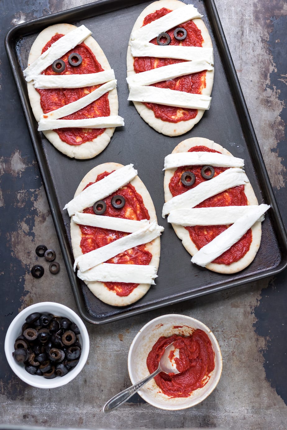 Pitas on a baking tray with pizza sauce and strips of mozzarella to look like mummies, with olives for eyes.