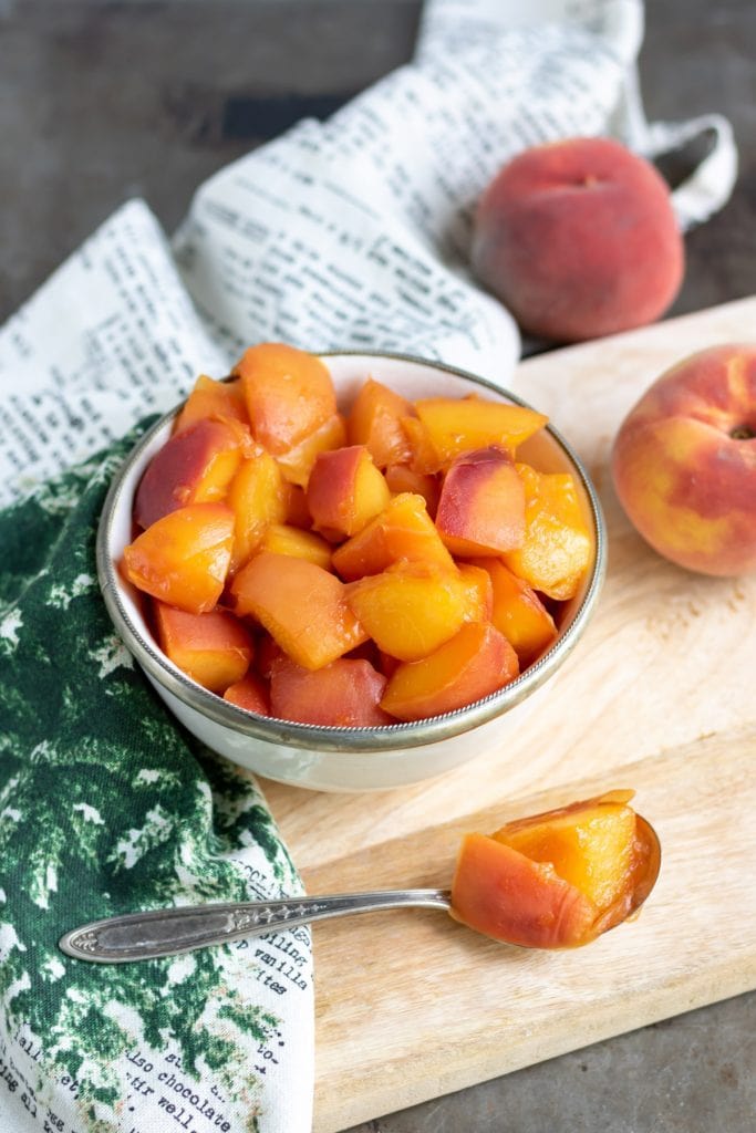Bowl of peach compote, with a spoonful in the front and peaches in the background.