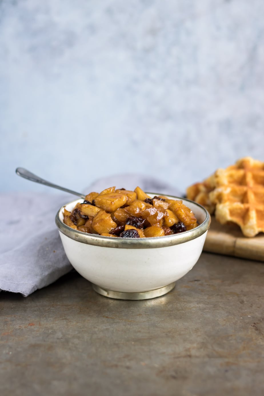 Bowl of apple compote with raisins and waffles in the background.