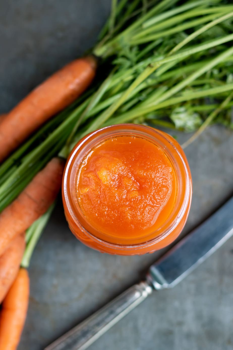 Looking down into a jar of carrot jam, next to a bunch of carrots and a knife.