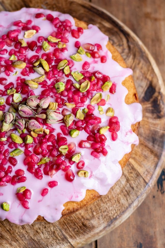 Looking down on a cake topped with pomegranate seeds, pistachios and rose petals.