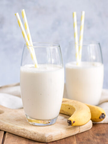Glasses of banana lassi with straws and bananas next to them.