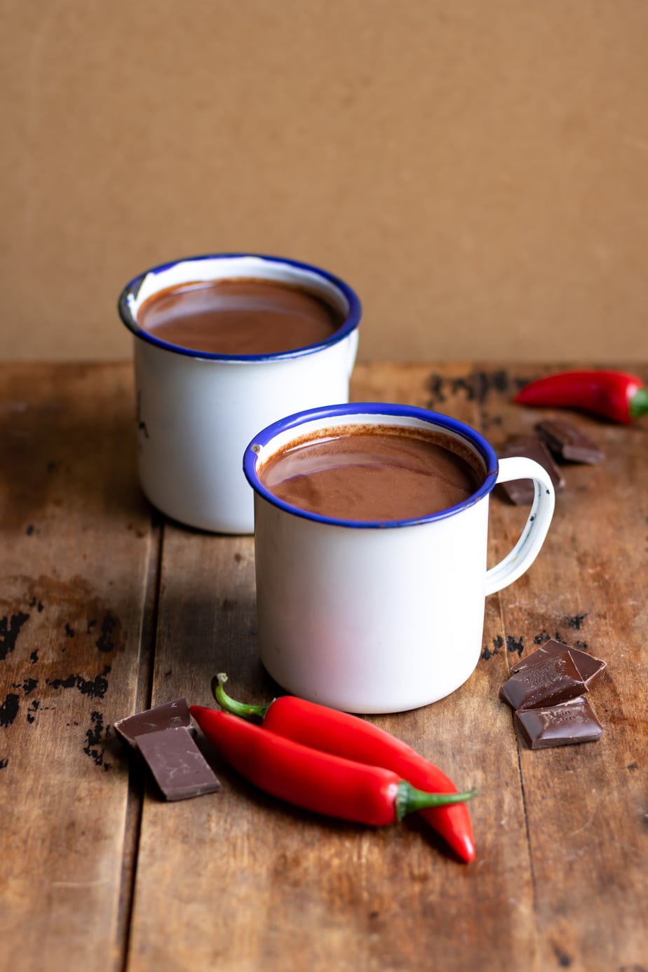 Table with two mugs of hot chocolate and chili peppers and chocolate on the table.