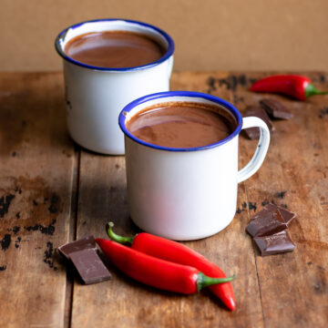 Table with two mugs of hot chocolate and chili peppers and chocolate nearby.