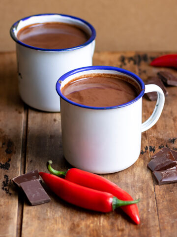 Table with two mugs of hot chocolate and chili peppers and chocolate nearby.