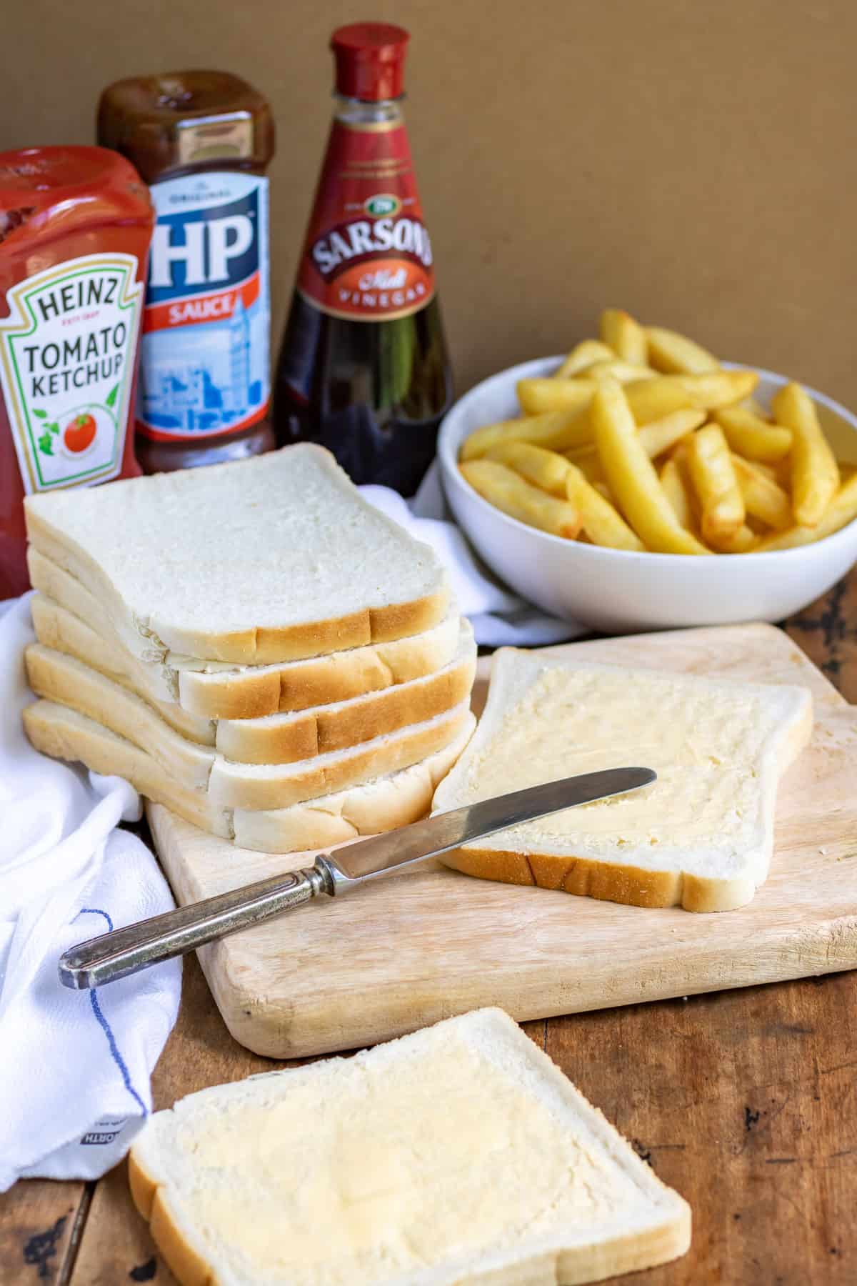 Stack of bread with a slice being buttered, in front of a bowl of fries.