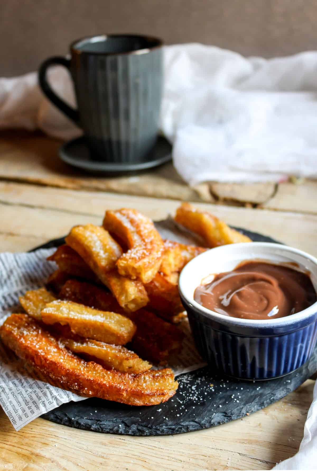 Table with a plate of churros and chocolate sauce, with a mug in the background.