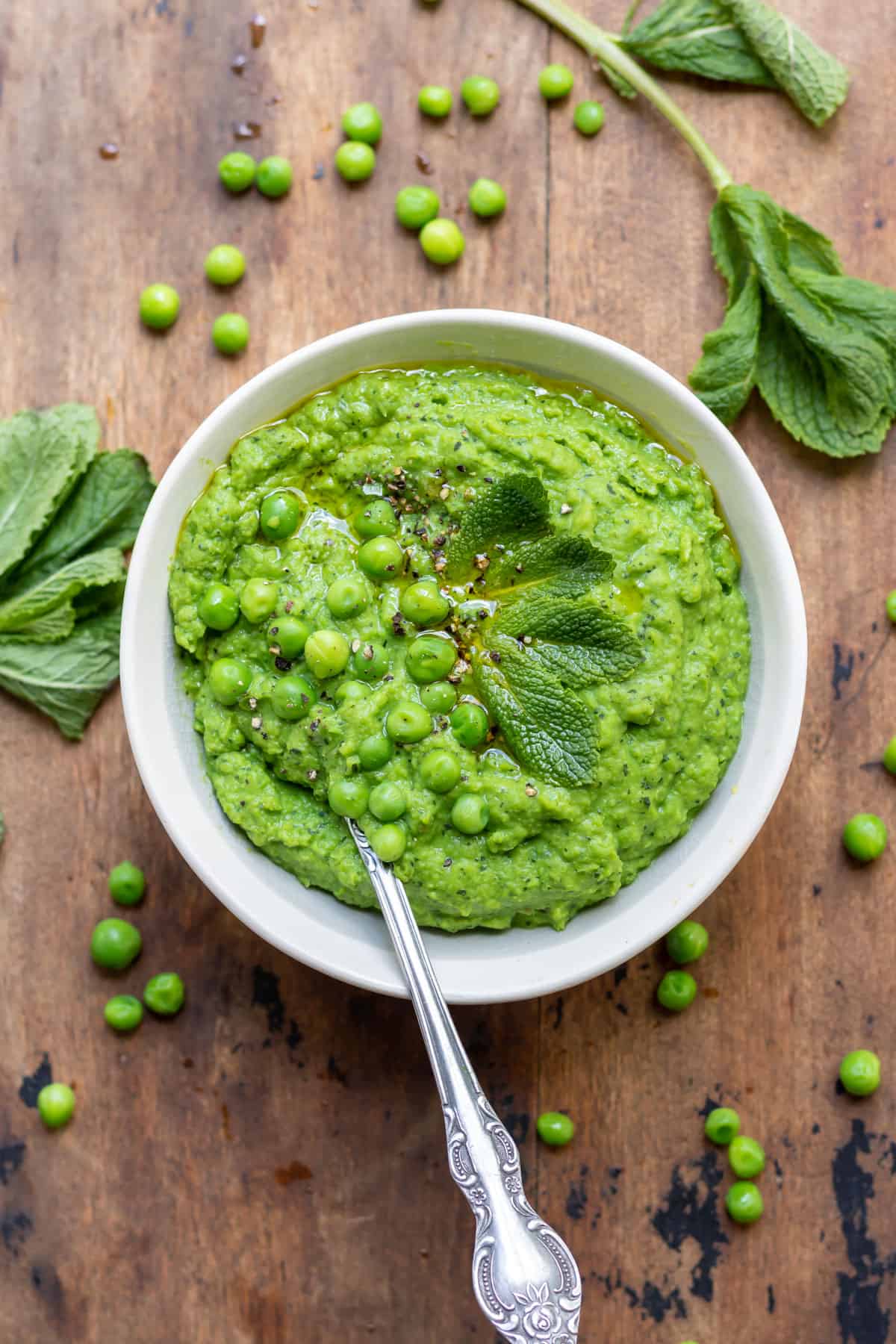 Wooden table with bowl of pureed peas, with peas and mint strewn around.