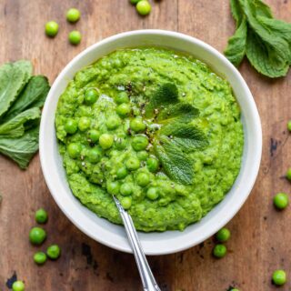 Wooden table with bowl of pureed peas, with peas and mint strewn around.