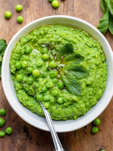Wooden table with bowl of pureed peas, with peas and mint strewn around.
