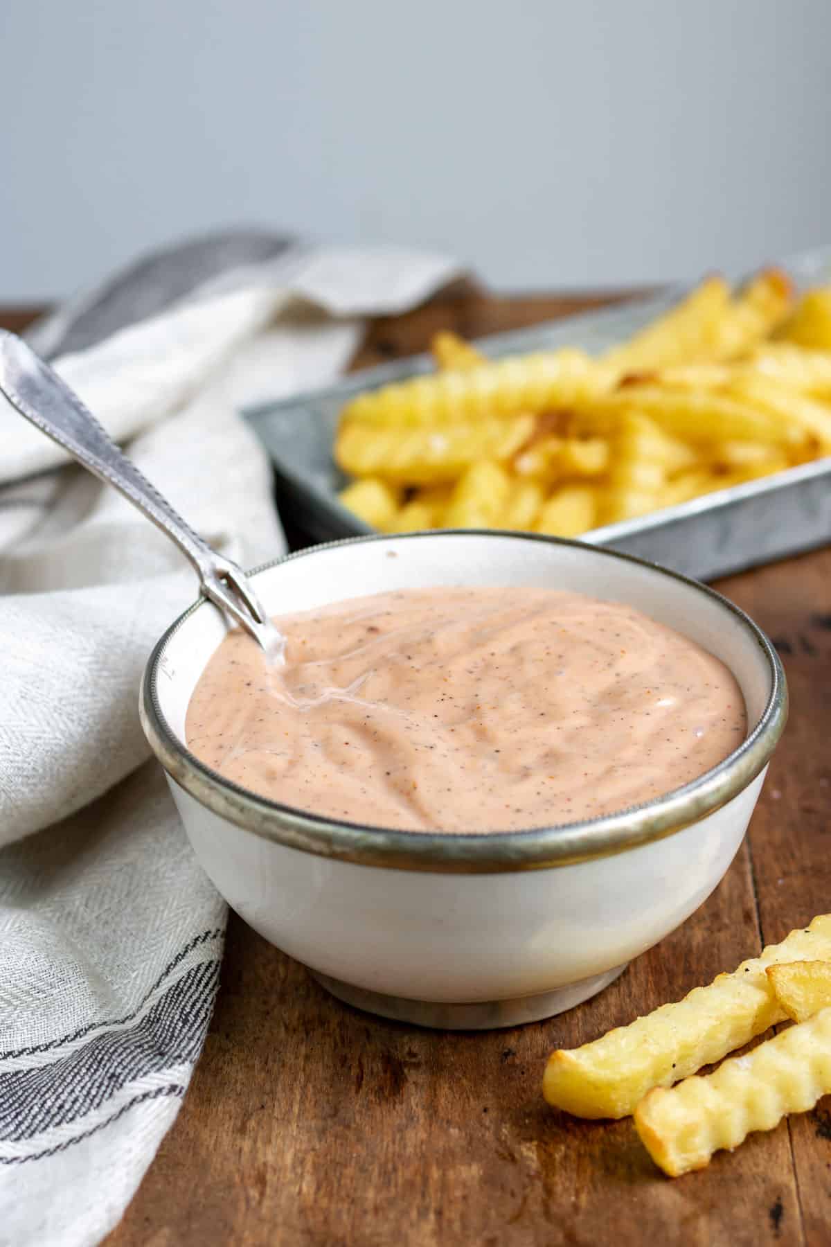 Spoon in a bowl of sauce in front of a dish of french fries.