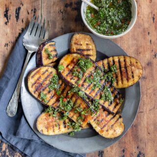 Wooden table with a plate of grilled eggplant steaks and a bowl of chimichurri.