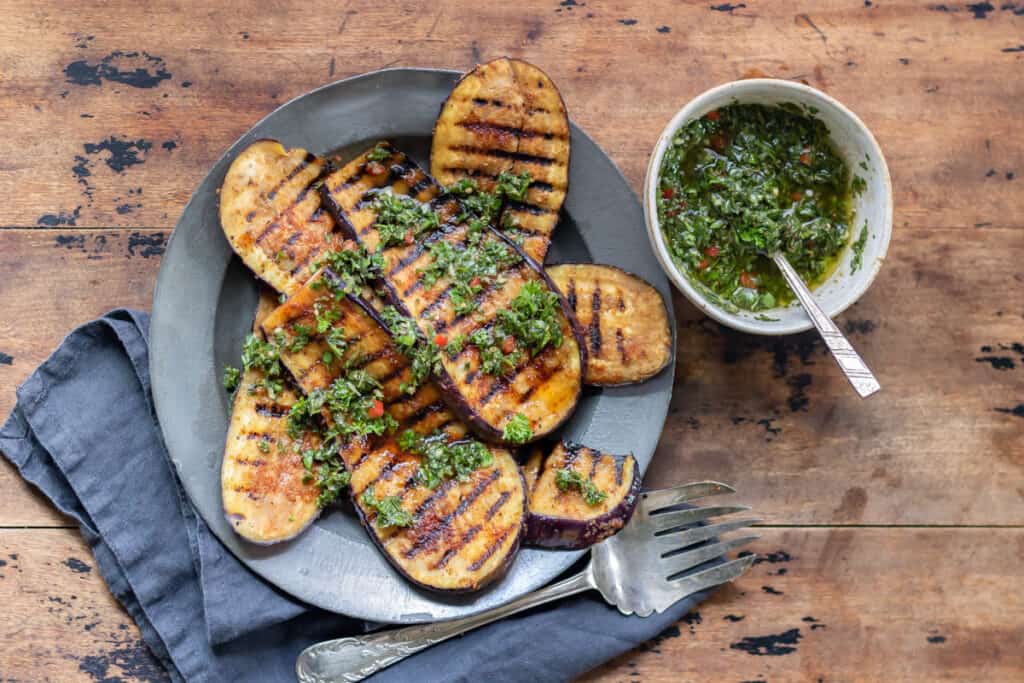 Wooden table with a plate of grilled eggplant steaks and a bowl of chimichurri.