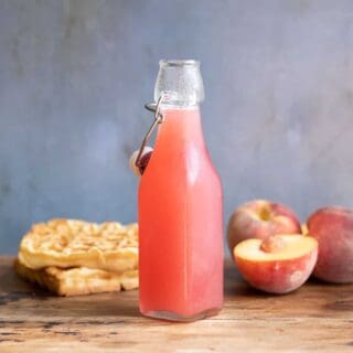 Bottle of fresh peach syrup next to peaches and waffles.