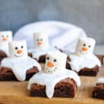 A wooden board with brownies topped with icing and marshmallows decorated to look like snowmen.