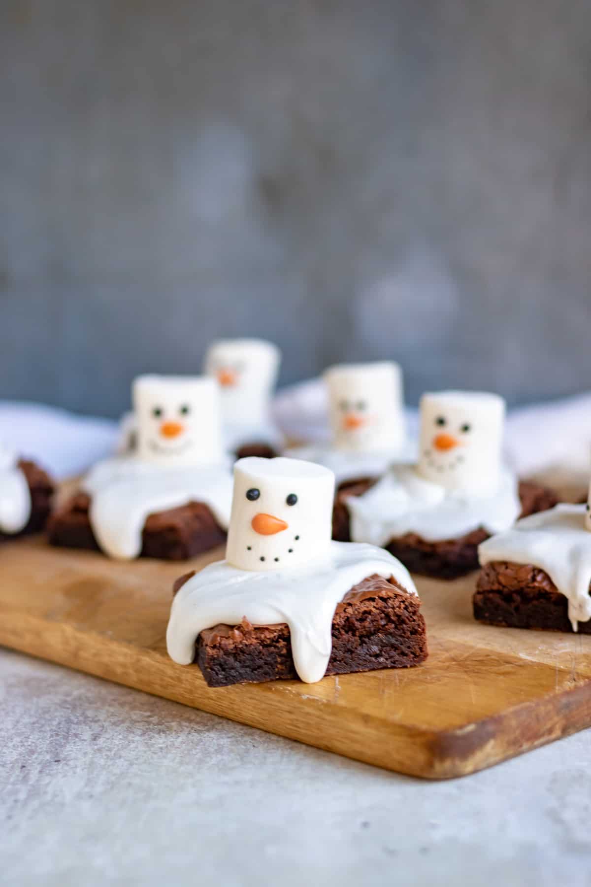 A wooden board with brownies topped with icing and marshmallows decorated to look like snowmen.