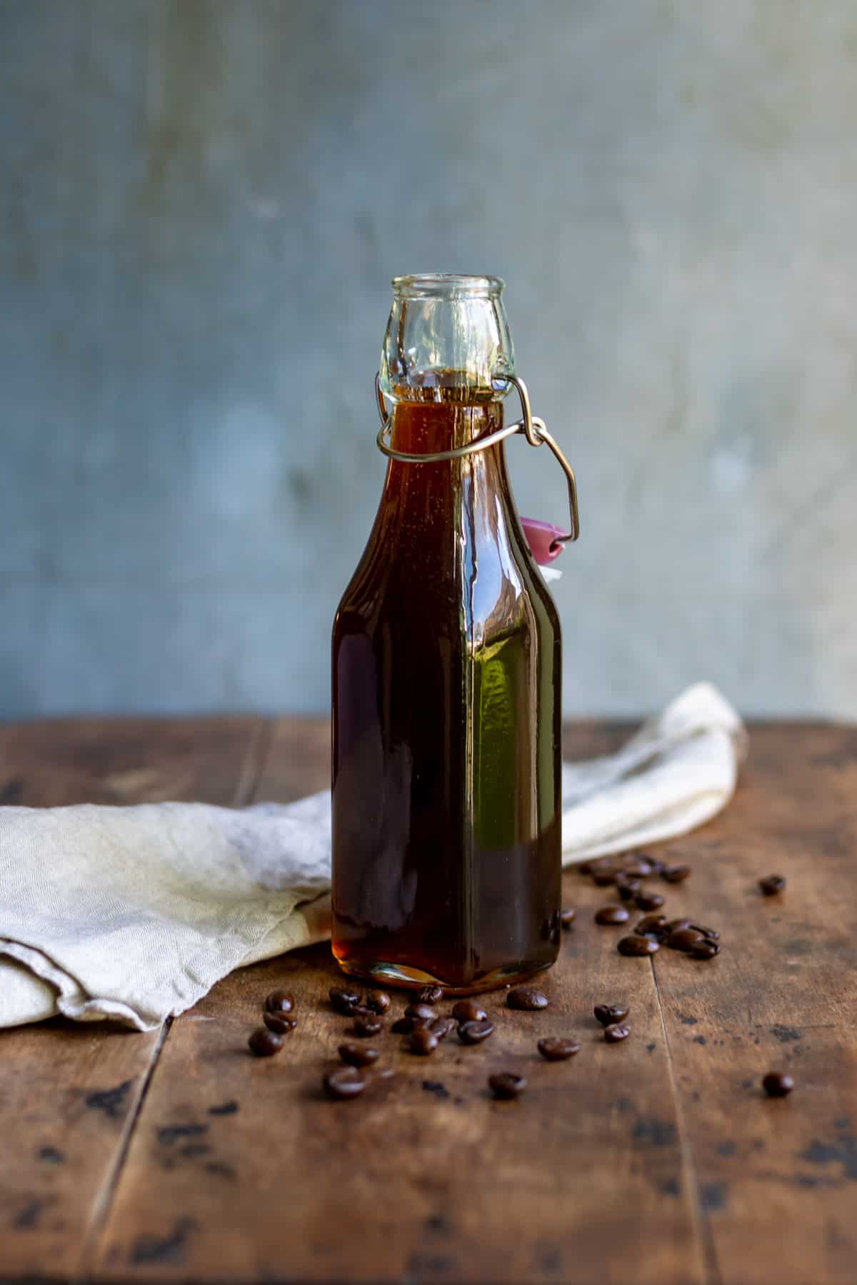 A wooden table with a glass bottle of coffee syrup with coffee beans scattered around.