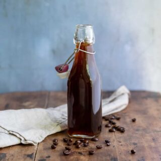 A wooden table with a glass bottle of coffee syrup with coffee beans scattered around.
