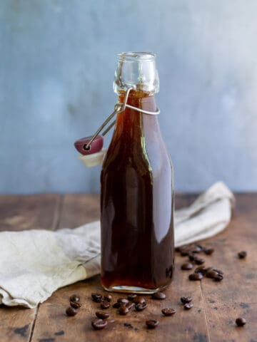 A wooden table with a glass bottle of coffee syrup with coffee beans scattered around.