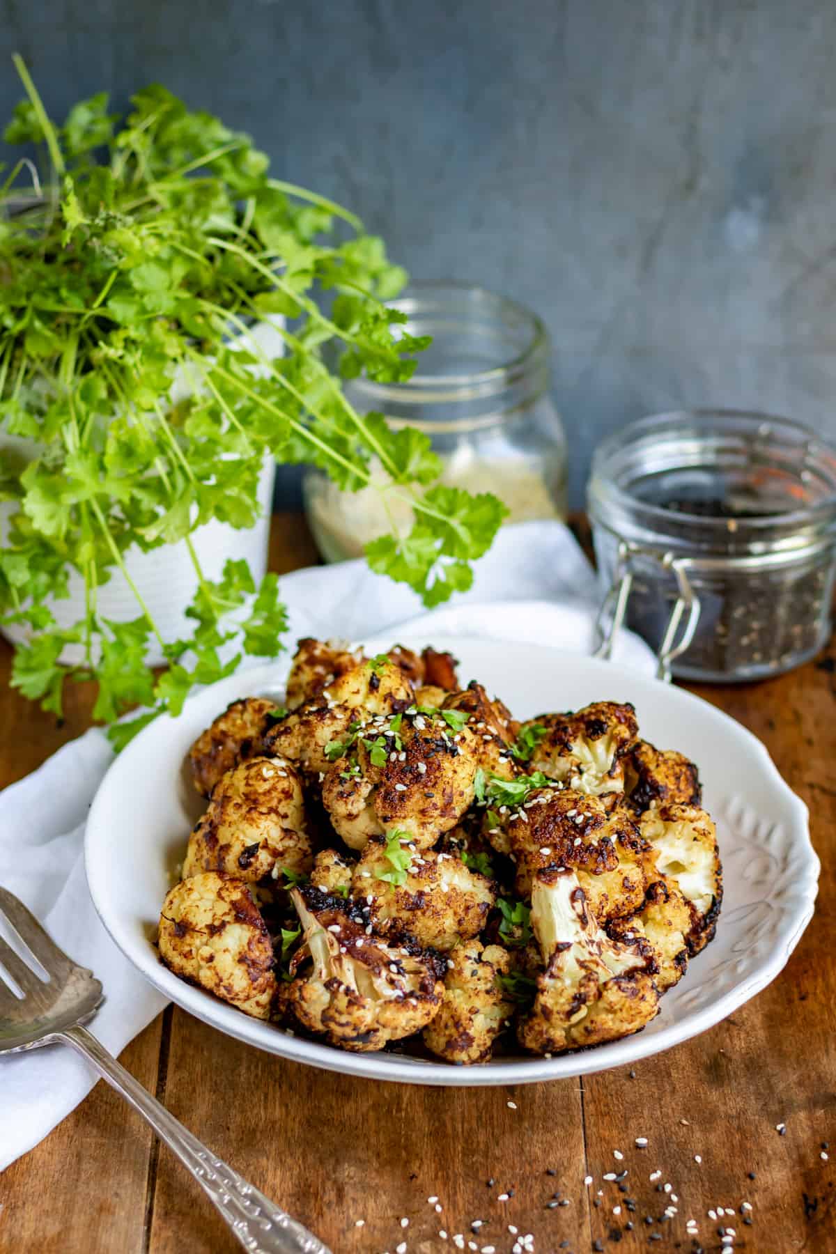 A table with a dish of roasted cauliflower, parsley and a jar of black sesame seeds.