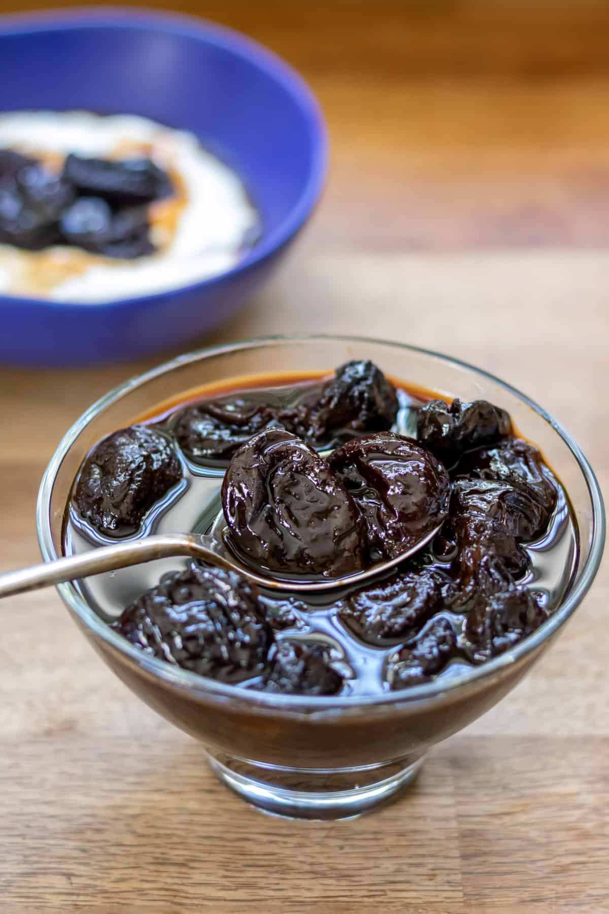 Spoon coming out of a dish of stewed prunes, with them over yogurt in a bowl in the background.