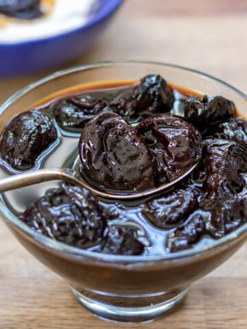Spoon coming out of a dish of stewed prunes, with them over yogurt in a bowl in the background.