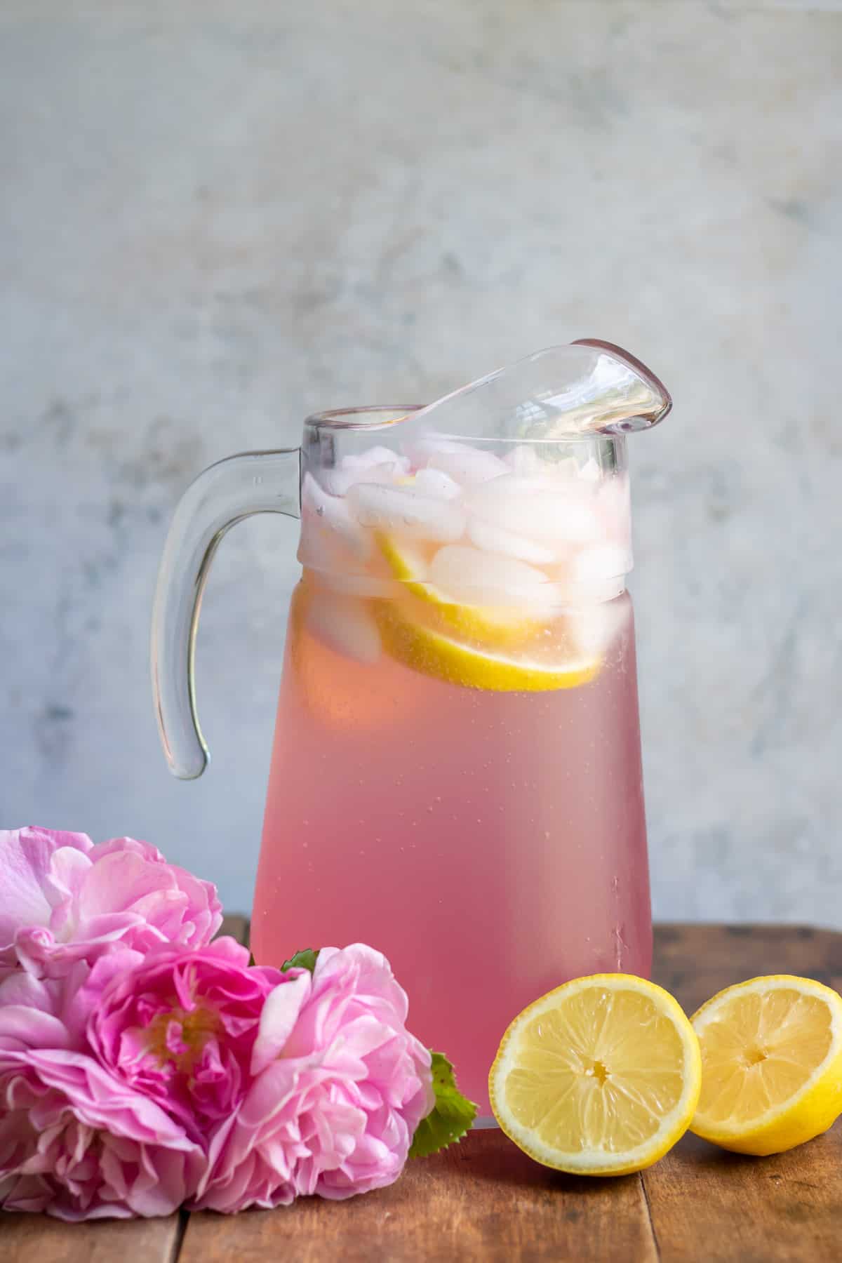 A table with a pitcher of lemonade with rose simple syrup, next to roses and lemons.