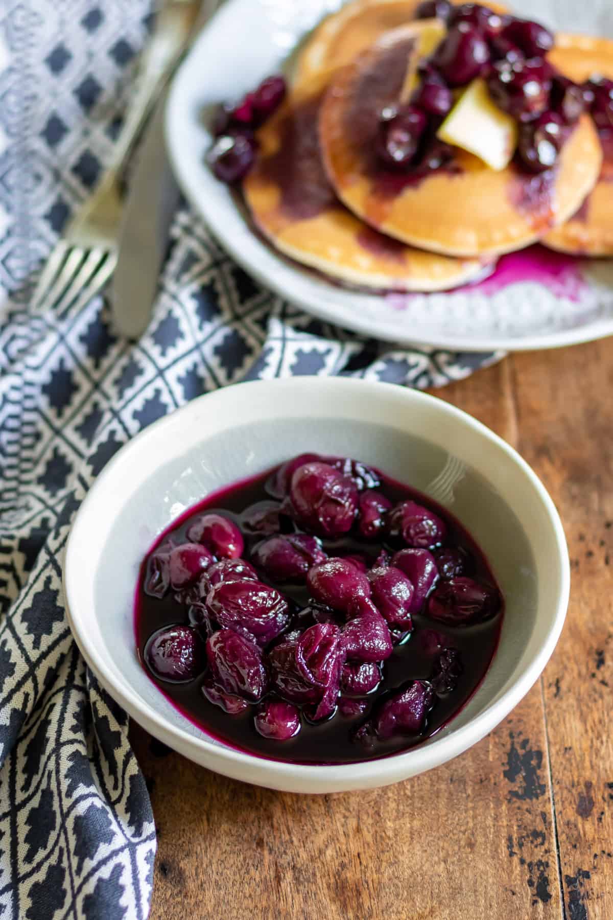 Dish of grape compote on a table with a stack of pancakes.