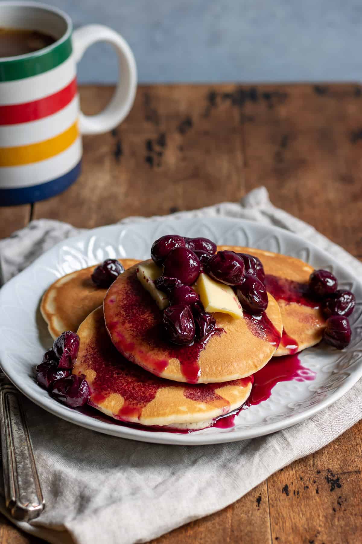 Table with a mug and plate of pancakes topped with grape compote.