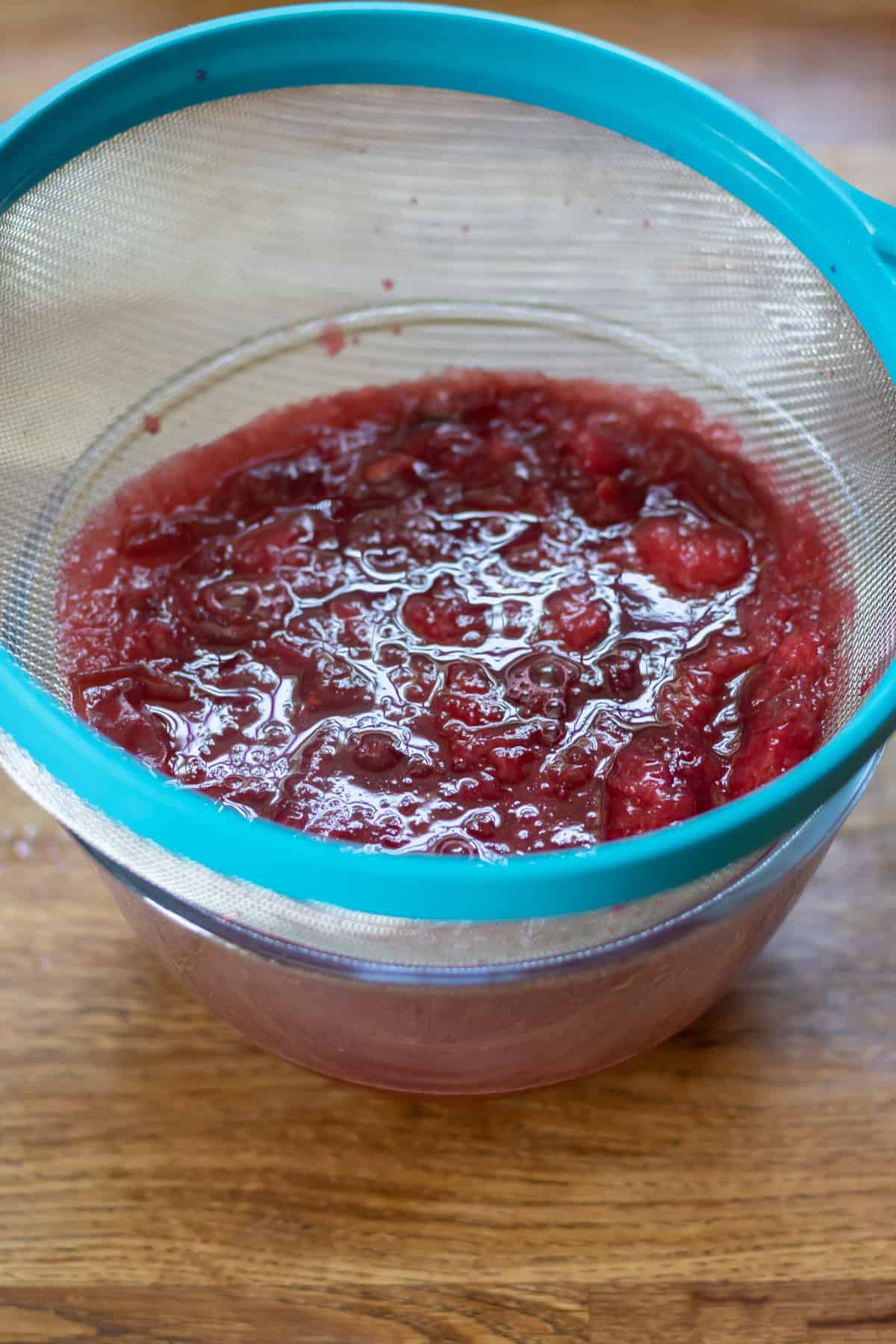 Straining the plum syrup into a bowl through a fine mesh strainer.