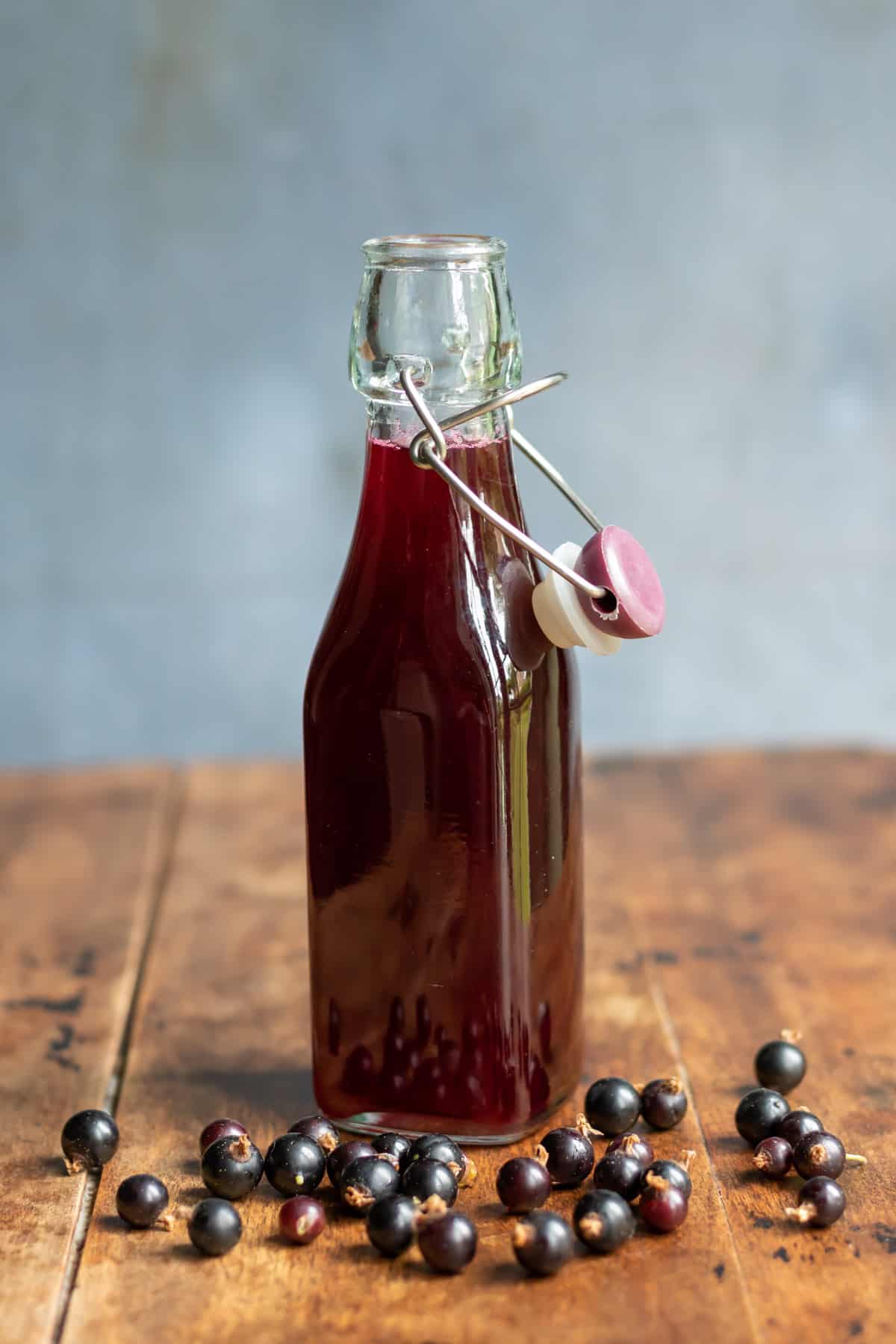 Wooden table with a bottle of blackcurrant simple syrup, with blackcurrants scattered next to it.