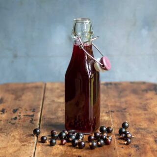 Wooden table with a bottle of blackcurrant simple syrup, with blackcurrants scattered next to it.
