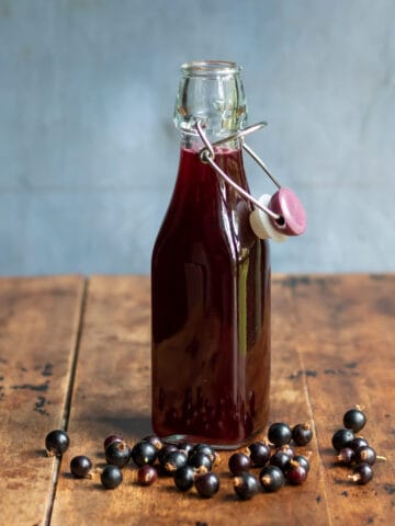 Wooden table with a bottle of blackcurrant simple syrup, with blackcurrants scattered next to it.