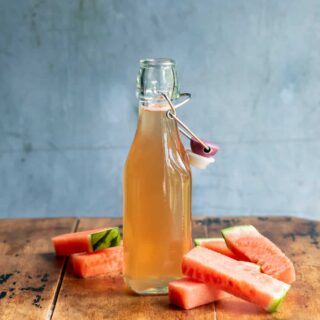 A bottle of watermelon simple syrup on a wooden table next to pieces of watermelon.