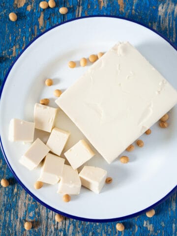 A block of silken tofu on a plate, next to soybeans and cut soft tofu.