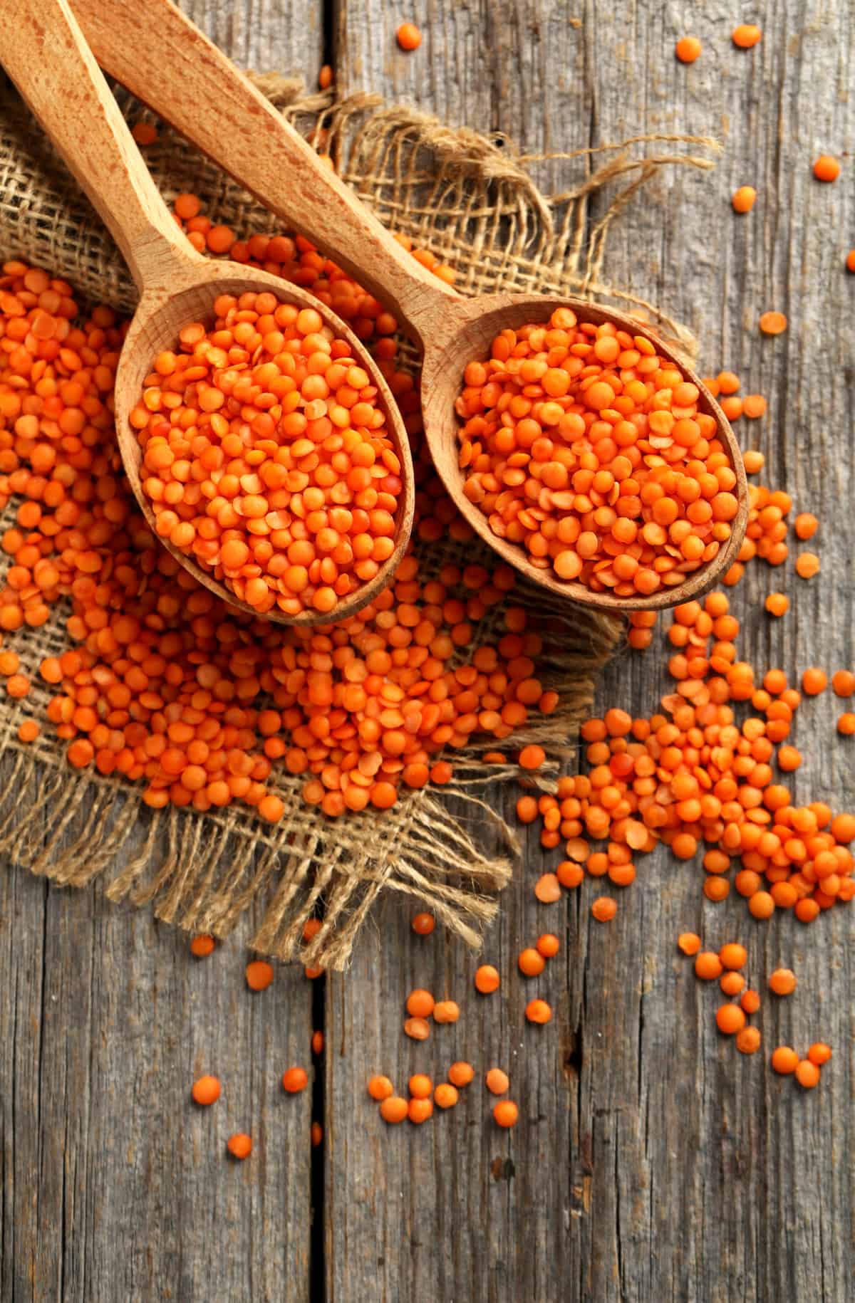 Two wooden spoons full of dried red lentils, with some spilled around on the wooden table.