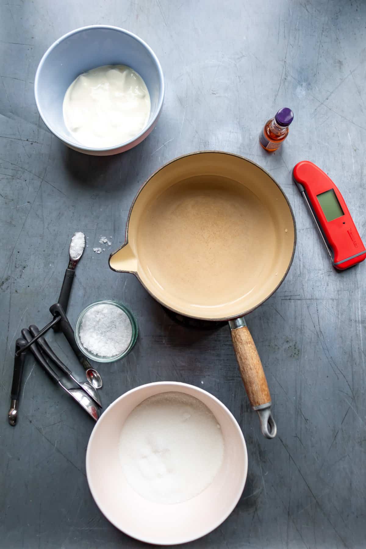 Pot, food thermometer, vanilla, sugar, cream, salt and measuring spoons on a table.