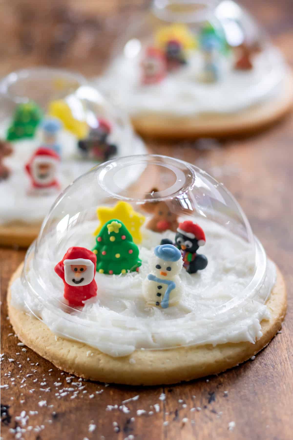 A wooden table with cookies topped with clear domes and filled with edible Christmas characters, so they look like Snow Globe Cookies.