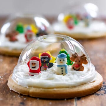 A wooden table with cookies topped with clear domes and filled with edible Christmas characters, so they look like Snow Globe Cookies.