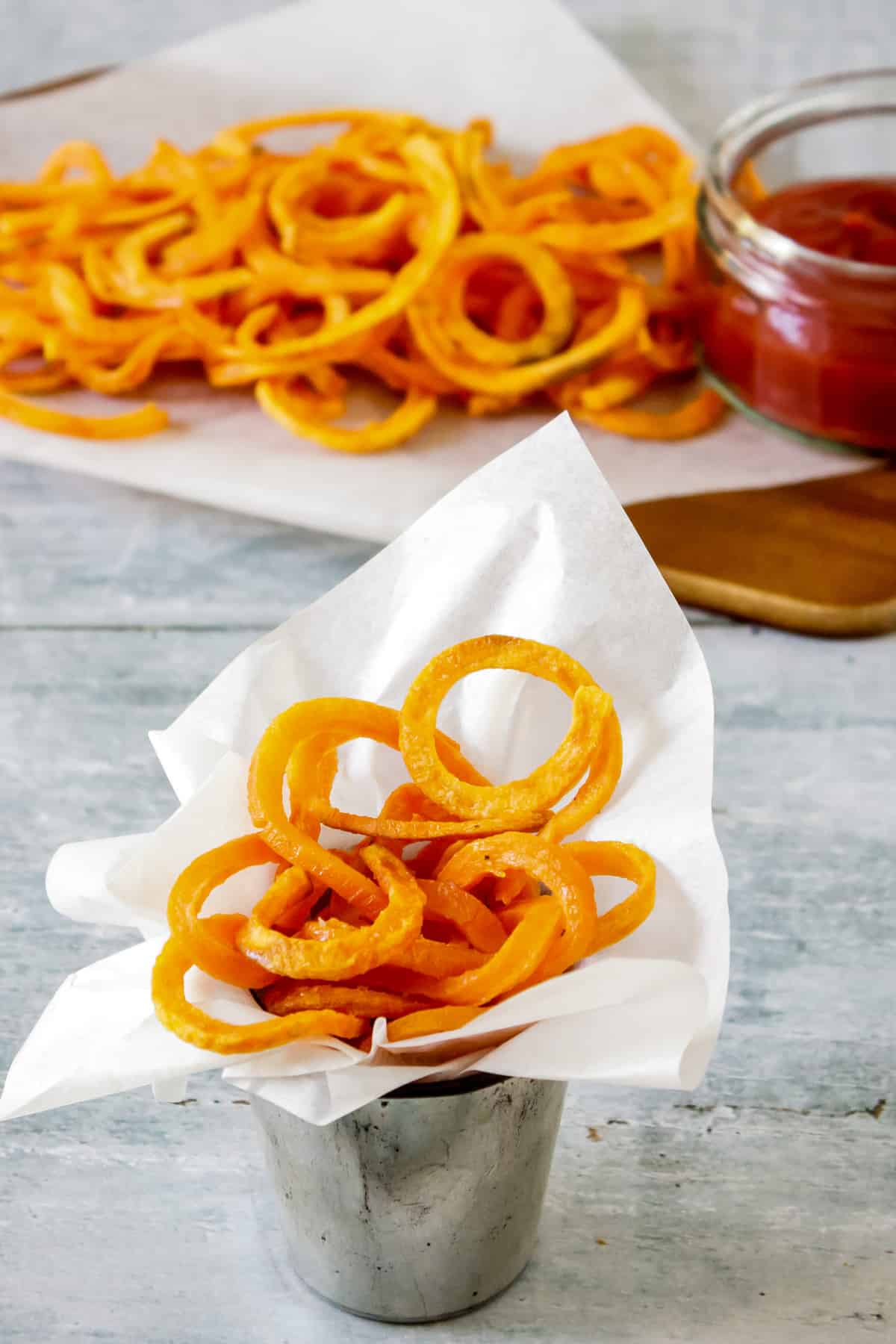 Table with a dish of spiralized sweet potato fries, plus a tray of them behind and a dish of ketchup.