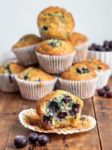 A table with a pile of buttermilk blueberry muffins, with one with a bite out in the front.